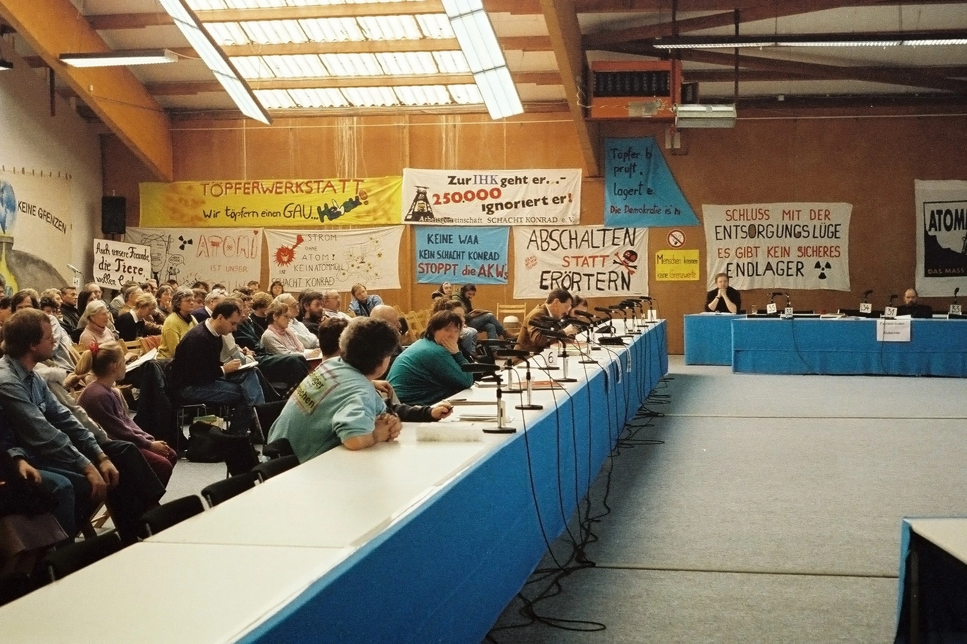 81 / 5.000 Übersetzungsergebnisse A group of people sit in a hall with protest placards in the background.