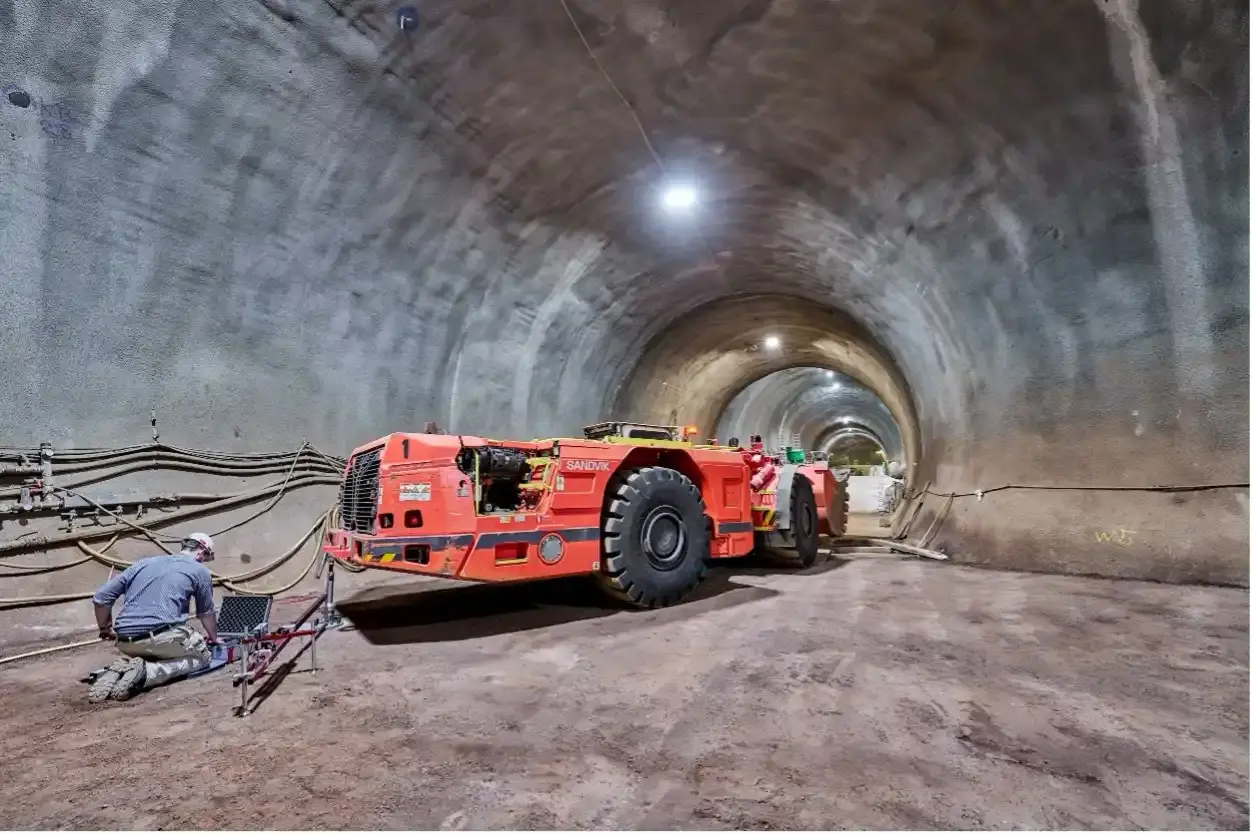 Ein orangefarbenes Kippfahrzeug steht in einem Tunnel eines Bergwerks. Hinter dem Fahrzeug kniet eine Person in Bergmannskleidung an einem geöffneten Koffer, der auf dem Boden steht.