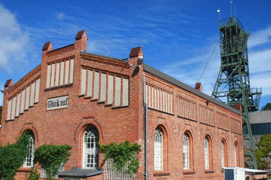Das Bild zeigt die Maschinenhalle der Schachtanlage Asse II. Das Gebäude besteht aus rotem Backstein. Im Hintergrund ist der Förderturm der Schachtanlage Asse II zu sehen. Dieser besteht aus eine grünen Stahlkonstruktion.